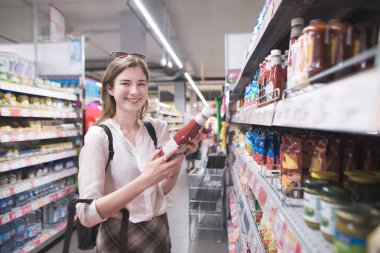 Happy, stylish girl standing in a ketchup bottle in the hands of a supermarket and looking into the camera. Happy buyer makes purchases in the supermarket. clipart