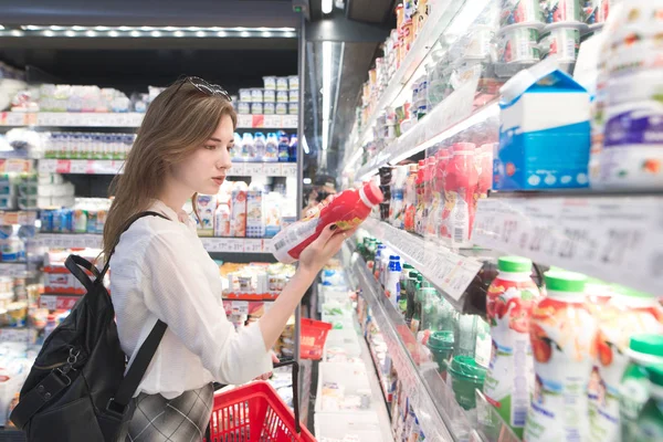 时尚迷人的女人站在冰箱与乳制品 持有酸奶在他的手中 并阅读标签 一个有吸引力的女孩在超级市场买酸奶 — 图库照片