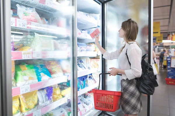 Stylish woman buys frozen food at a supermarket. Attractive young woman stands beside a freezer and chooses frozen foods. Purchase of products in a supermarket.