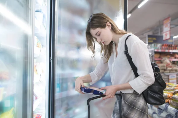 Woman is in the supermarket near the refrigerator and looks at the frozen foods in her hands. Girl chooses and buys frozen food. Purchase of products in a supermarket