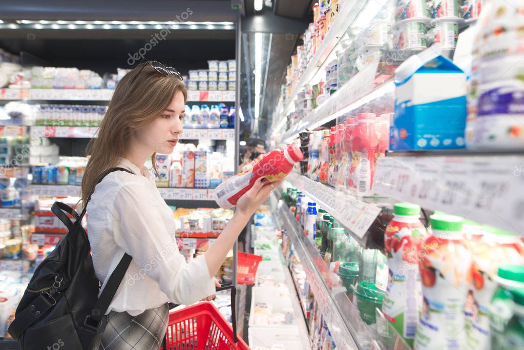 Stylish attractive woman is standing by the refrigerator with dairy products, holds yogurt in his hands and reads the label. An attractive girl buys a yogurt in a supermarket.
