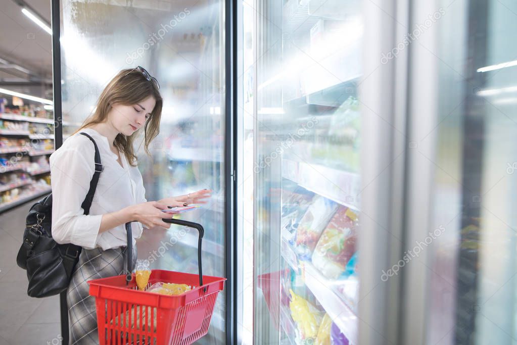 A young girl with a red basket is standing by the refrigerator and chooses frozen dumplings. Choosing and buying frozen foods at a supermarket.