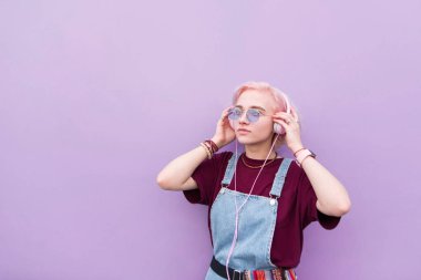 Pembe saçları ve güneş gözlüğü ile şık kız mor bir arka plan üzerinde kulaklık müzik getiriyor. Genç kız pembe kulaklık müzik dinlemek portresi.