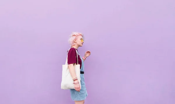 时尚的女孩走在蓝色的墙壁与购物袋 粉红色的头发 眼镜和购物的女孩在紫色的墙壁背景 — 图库照片