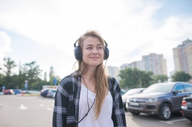 Park yerinde duran ve kulaklık müzik rahat kıyafetler içinde bir kız sokak portresi. Kentsel peyzaj zemin karşı Kulaklığımda gülümseyen kız.