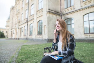 Bir üniversite kampüsünde bir bankta kitaplar ile onun kucağına oturmuş, telefonda konuşurken ve gülümseyen pozitif öğrenci kız. Güzel kız öğrenci üniversite yakınında sokakta oturan ve dinlenme