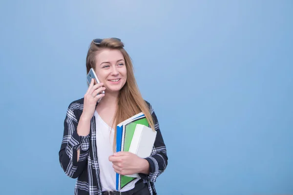 微笑的女孩在电话里聊天 拿着笔记本和蓝色背景的书 快乐的学生在智能手机上交谈 看着远方 孤立的蓝色背景 Copyspace — 图库照片