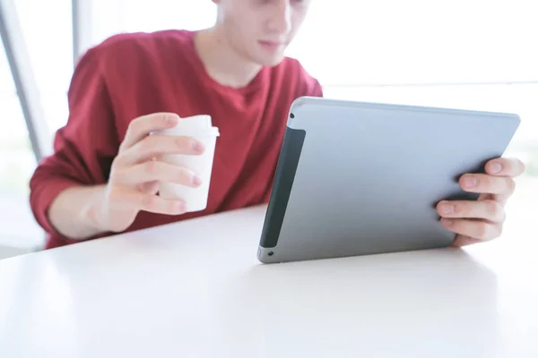 タブレットおよびコーヒーと手のガラスが付いて赤いトレーナーのテーブルに座っての若い男 カフェでタブレットを持つ学生 フォーカスとクローズ アップをタブレットします タブレット上のビデオを見る — ストック写真