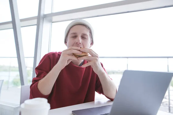 カジュアルな服装の若い男とファーストフードのお店でノート座ってハンバーガーを食べる 学生は サンドイッチを刺さ 仕事とファーストフード カフェで食事 — ストック写真