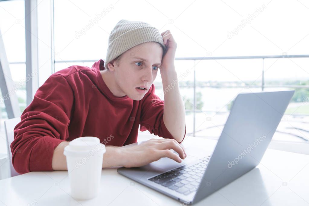 Young man is a freelancer in casual clothing, he concentrates on a laptop in a cafe. A stylish young man looks at the laptop screen with amazement. Work on a laptop in a cafe