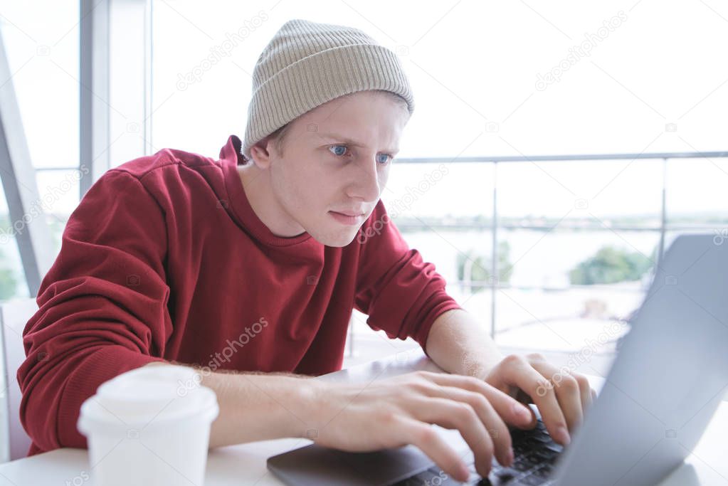 Serious young man in casual clothing, sitting at the table and typing the text on the keyboard of the laptop.Young freelancer works on a laptop in a light cafe.Blogger writes an article on a computer