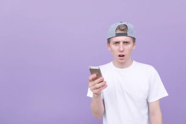 Genç adam bir beyaz T-shirt ile bir akıllı telefon elinde mor bir arka plan üzerinde duruyor ve kameraya görünüyor hayran. Mor bir arka plan bir smartphone kullanarak bir öğrenci portresi.