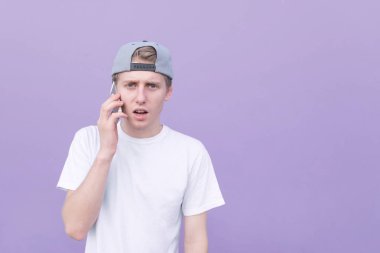 Mor duvar arka plan üzerinde konuşmak ve kameraya bakarak beyaz bir T-shirt bir duygusal genç adam portresi. Genç öğrenci mor bir arka plan telefonda konuşurken