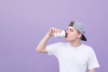 Komik öğrenci bir beyaz T-shirt ve mor bir arka plan ve fotoğraf makinesine bir göz bakarak bir bardaktan kahve içmek kap. Portre arka planını mor bir duvar bir genç adam içme kahve