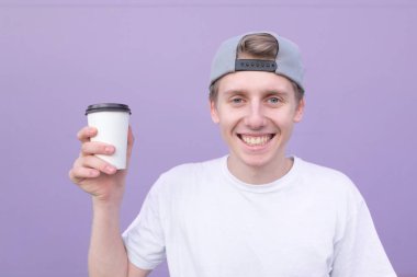 Bir fincan kahve bir mor duvar arka planı ile ayakta, kamera seyir ve gülümseyen bir beyaz T-shirt giyen mutlu genç adam. Bir fincan kahve mor bir arka plan ile gülümseyen öğrenci