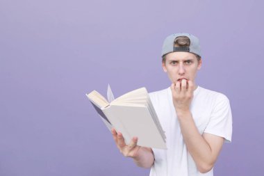 Beyaz bir T-shirt düşünceli genç adam ve onun elinde bir kitap bir mor duvar arka plan üzerinde duruyor ve kameraya görünüyor. Mor bir arka plan üzerinde elinde bir kitapla duygusal öğrenci.