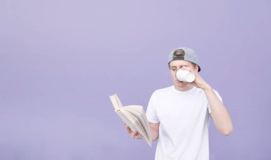 Bir kap ve bir kitap okur ve pastel bir mor arka plan üzerinde kahve içiyor beyaz tişört genç bir adam portresi. Öğrenci bir kitap okur ve mor duvar arka plan üzerinde kahve içer.