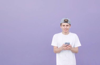 Adam stand Mor pastel arka plan üzerinde elinde bir akıllı telefon ile gülümseyen, kulaklık müzik dinler ve kameraya benziyor. Sokak mor bir duvar bir arka plan üzerinde genç bir adam portresi.