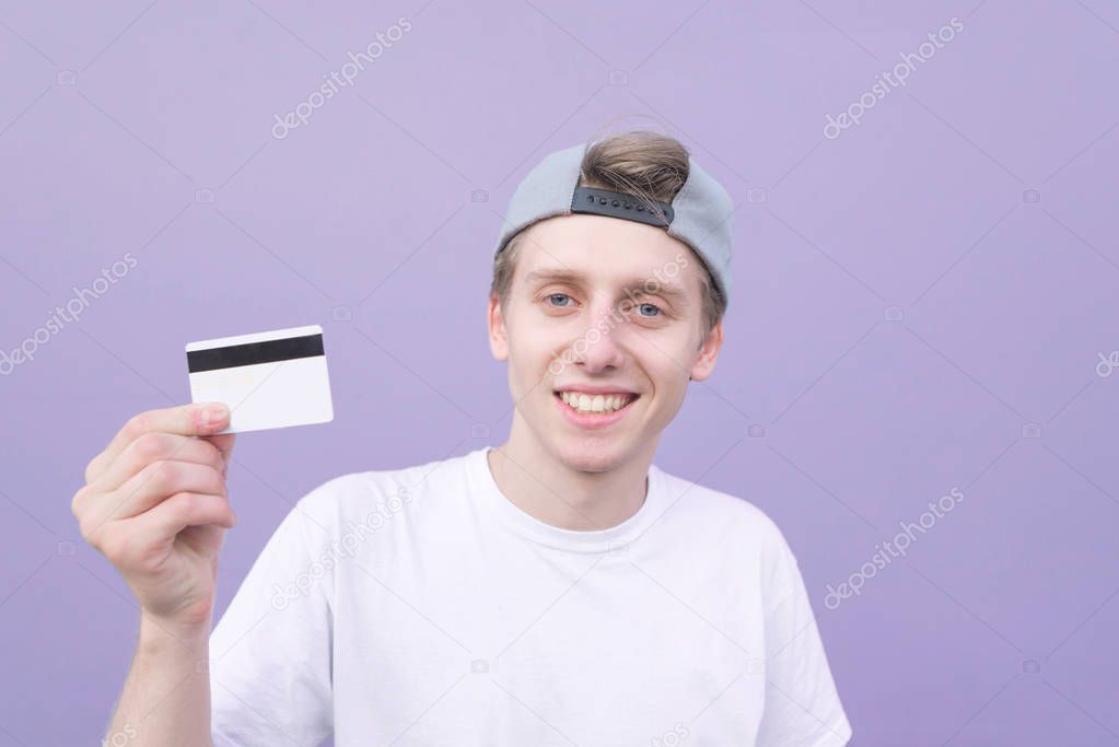 Happy young man in a white T-shirt holds a bank card in his hands and smiles on a purple background. Smiling student with a credit card on a purple background