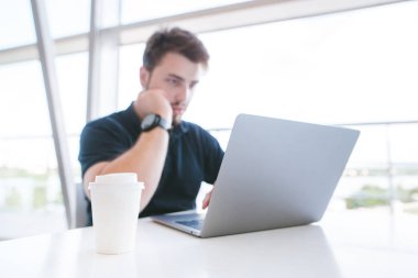 Beyaz cam pencerenin yakınındaki bir dizüstü bilgisayar üzerinde çalışan bir adam arka plan üzerinde kahve. Bir sakallı bir kafede oturur ve bir dizüstü bilgisayar kullanarak.