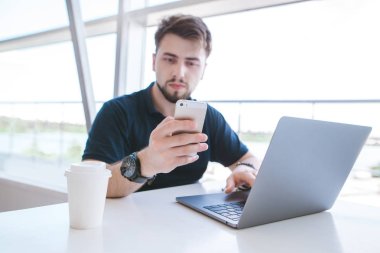 Çekici adam sakalı ve bir siyah T-shirt bir dizüstü bilgisayarda çalışırken bir akıllı telefon kullanır. Bir bardak kahve ile hafif bir kafede çalışıyorum. Belgili tanımlık smartphone üzerinde odaklanmak.