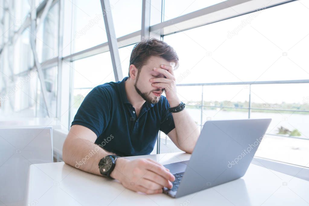 Businessman works on a notebook in a cafe and makes face palm. Sad man in casual clothes is sitting at the table and closes his face with his hand.