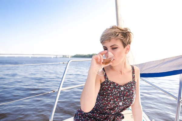 Beautiful woman in her dress sitting on a yacht sails and drinking wine from a glass. Portrait of a relaxing woman, floating by sea on a yacht on a summer sunny day. Gairl drinks alcohol on a yacht