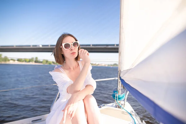 Portrait of an attractive woman sitting on a sailing yacht and looking at camera at the background of the river and bridge. Girl is resting on a yacht on a sunny, warm day