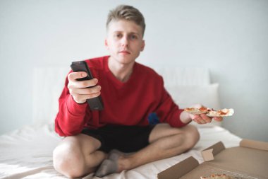 Bir parça pizza ve onun elinde bir uzaktan kumanda ile rahat bir parlak oda oturan ve televizyon izlerken şık genç adam. Pizza ve tv ile hafta sonu. Uzaktan kumanda ve bir parça pizza odaklanmak