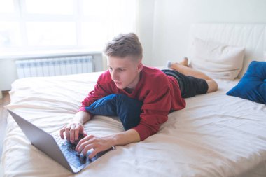 Konsantre genç adam evde yatakta yatıyor ve bir dizüstü bilgisayar kullanarak. Serbest meslek sahibi bir laptop evde rahat bir oda içinde çalışır. Internet üzerinde çalışma.