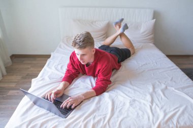 Konsantre genç adam bir yatakta rahat parlak oda içinde koyar ve bir dizüstü bilgisayar Internet üzerinde çalışır. Evde Internet üzerinde çalışıyorum. Öğrenci bir dizüstü bilgisayar yatakta kullanır.