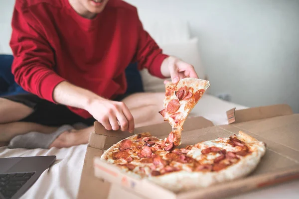 赤いトレーナー若い男は 自宅のベッドの上に座っているし ボックスからピザの食欲をそそる作品がかかります 男性の手は ピザの美味しい部分を取る ティーンエイ ジャーは ピザの部分を食べるつもりです — ストック写真