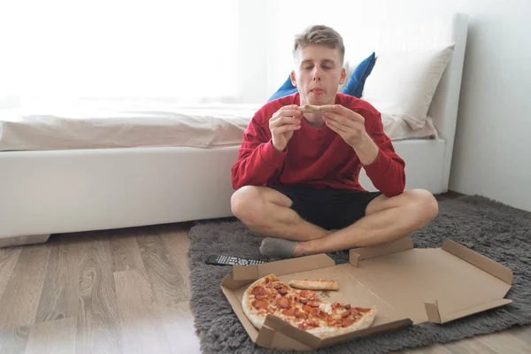 赤いトレーナーに魅力的な若者階の彼の部屋に座っているボックスからピザを食べる 喜びを取得します ランチ ピザ配信 週末に自宅でおいしいピザを食べる — ストック写真