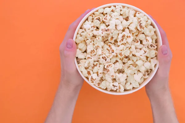 Men\'s hands hold a paper cup with popcorn on an orange background. Man with popcorn in his hands.solated. Cinema Concept. Flat lay. Copyspace.