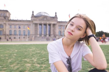 Reystag Arka çimenlikte oturan ve gülümseyerek kameraya bakarak çekici bir kız portresi. Avrupa'nın sevimli turist kız pozlar tarihi mimari arka planı