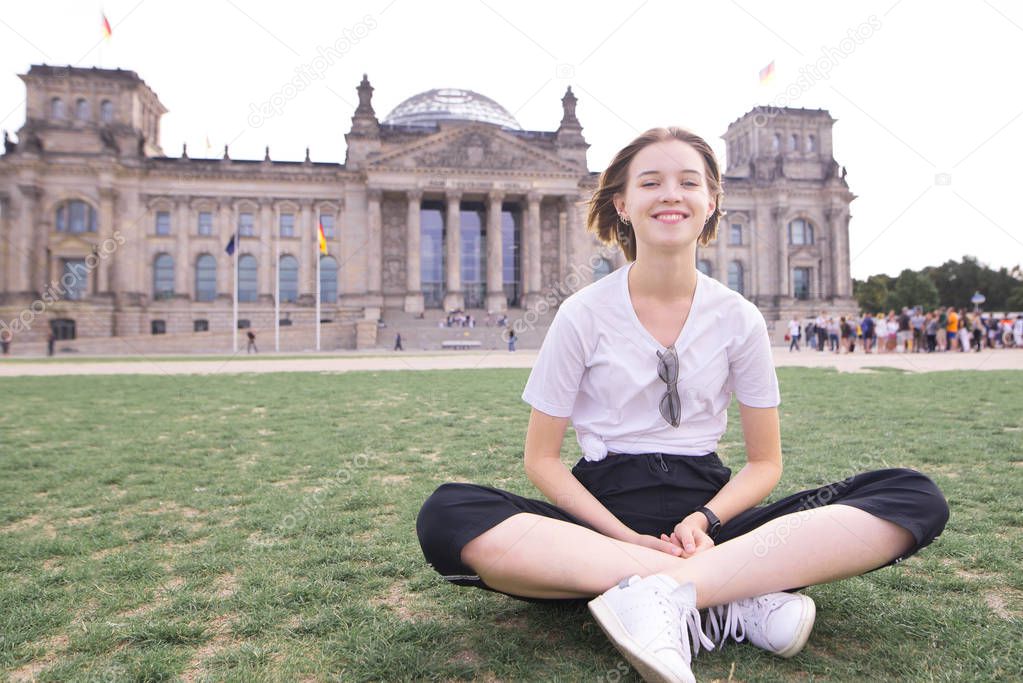 Smiling happy girl in a white T-shirt sits on the lawn on the background of the Reichstag, Berlin, Germany. Attractive girl traveler in Berlin