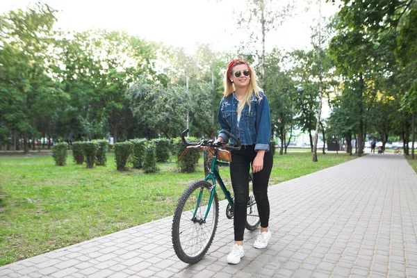 年轻快乐的女孩穿着时髦的衣服 在公园里走着绿色的自行车 微笑着 傍晚骑自行车沿小巷在公园 — 图库照片
