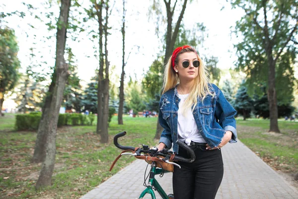 Portrait of attractive girl in park with bicycle while walking. Young woman with a bike stands in the park. Active leisure with cycling.
