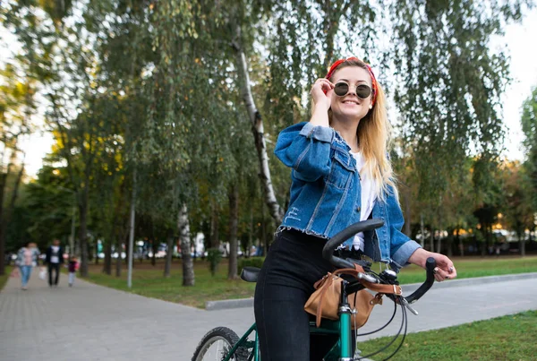 Happy, stylish girl with a bicycle standing in the park looking away and smiling. Attractive woman rides a bicycle in the park. Walk on the bike