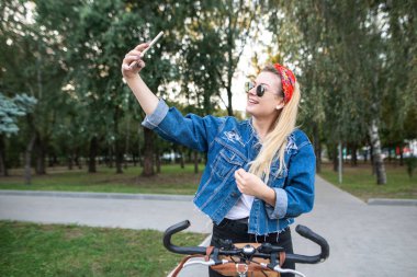 Mutlu şık kız gülümseyerek ve fotoğraf makinesi smartphone vasıl seyir parkta bisiklet ile selfie yapıyor. Selfie akıllı telefon yapmak için bir bisiklet sürme şık genç kadın