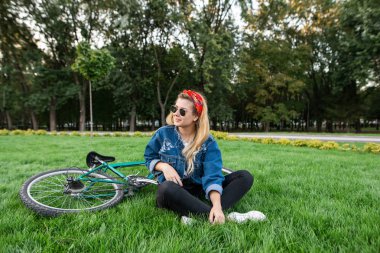Stylish attractive young woman sits on a lawn in a park on a lawn and smiles. Beautiful girl in a stylish outfit resting on the grass while walking on a bike.Relax outdoors clipart