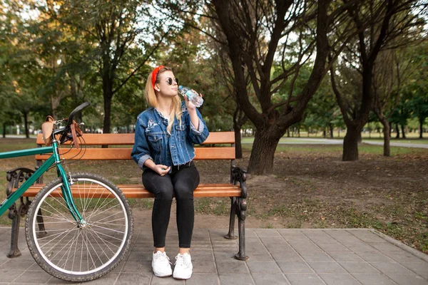 魅力的なスタイリッシュな女の子が彼女の手に水のボトルと自転車の近くの公園のベンチに座っています 公園で ボトルから水を飲んでベンチで休んで女性 — ストック写真