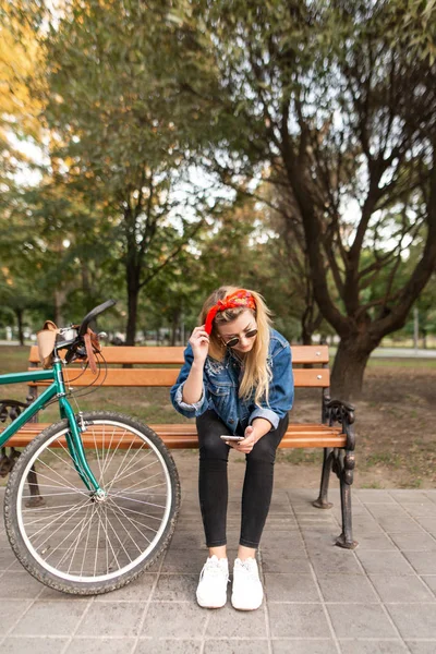 自転車に乗って 公園のベンチに座っていると スマート フォンを使ってスタイリッシュな女の子 女性は 公園内を歩きながらスマート フォンでインターネット接続を使用してください — ストック写真