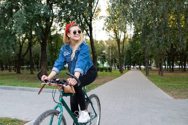 スタイリッシュな服とサングラスでスタイリッシュな幸せな女の子公園 横に見える笑顔で自転車に乗る 公園で自転車の上を歩きます 概念の残りの部分で活躍 — ストック写真