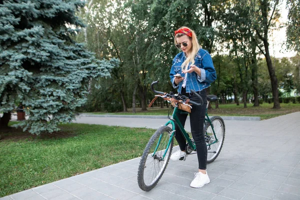 スタイリッシュな魅力的な女性は自転車に乗って公園に立っているし スマート フォンを使用します スマート フォン上女の子の自転車ルートを見る — ストック写真