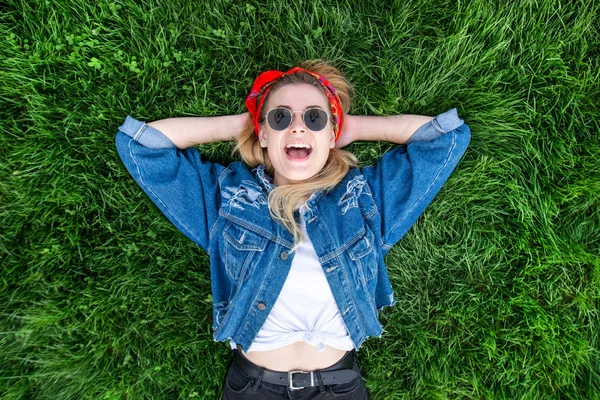 Emotional stylish woman in bright clothes lays on the grass, looks from above, looks into the camera and smiles. Happy emotional girl in stylish clothes lays on the lawn. Top view