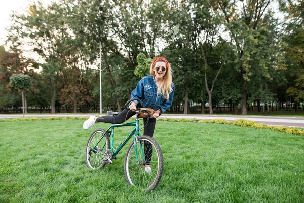 幸せな女の子は カメラと笑顔を見て緑の芝生公園内で自転車に座っています スタイリッシュな服でスタイリッシュな幸せな女公園と笑顔で自転車に乗る 自転車の上を歩く — ストック写真