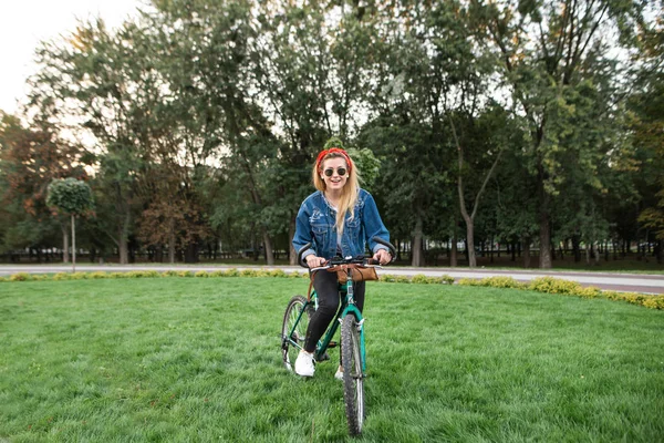 Stylish hipster girl rides a bicycle on a green lawn in the park looking at the camera and smiling. Walk on the bike in the park. Active rest on a bicycle. Stylish young woman riding a bike