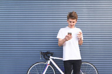 Portre genç bisikletçi duvar arka plan üzerinde. Bir fincan kahve ayakta duvara yakın bir bisiklet arka plan üzerinde olan yakışıklı genç adam bir smartphone kullanarak. Boşaltmak