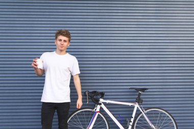 Yakışıklı genç bir beyaz T-shirt bir mavi duvar arka planı elinde kahve ile bir bisikletle duruyor. Kamera ve gülümsüyor arıyorum. Duvar. Boşaltmak
